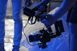 Hands holding a professional video camera rig, with focus on the equipment in a dimly lit setting. The background features blurred white and blue tones, creating a cinematic environment.