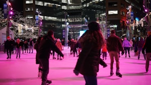 People are enjoying ice skating at an outdoor rink surrounded by tall buildings adorned with decorative lights. The rink is lively with skaters in winter attire moving in different directions, and the area is illuminated with purple and white lights, contributing to a festive atmosphere.