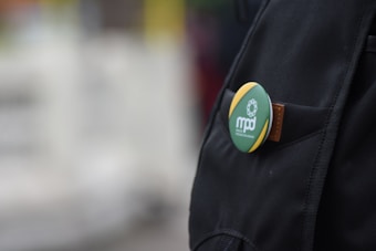 A close-up view of a black backpack featuring a decorative round pin button with green, yellow, and black colors. The button has a white floral design and the text 'mpd' on it, attached to the backpack's pocket or strap.