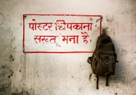 A weathered, white wall with a prominent message written in bold red Hindi text, surrounded by a red rectangular border. A backpack is hanging on a hook to the right side of the text. The wall has a rough texture with visible marks and scratches.