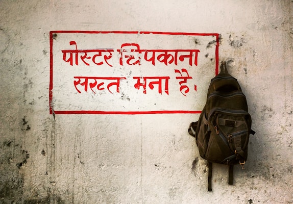 A weathered, white wall with a prominent message written in bold red Hindi text, surrounded by a red rectangular border. A backpack is hanging on a hook to the right side of the text. The wall has a rough texture with visible marks and scratches.