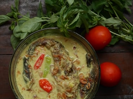 A bowl of creamy soup featuring sliced peppers and garnished with herbs and fried onions. Two fresh tomatoes rest beside it, accompanied by a bunch of fresh green leaves on a wooden surface.