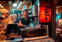 A busy and lively small Japanese restaurant kitchen with a chef grilling food over charcoal. The scene is warmly lit, and several patrons are visible enjoying their meals at the counter. The ambiance is cozy, with a red paper lantern providing ambient lighting.