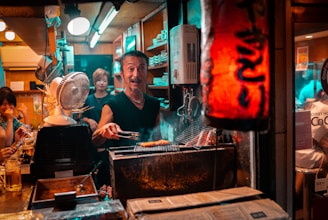 A busy and lively small Japanese restaurant kitchen with a chef grilling food over charcoal. The scene is warmly lit, and several patrons are visible enjoying their meals at the counter. The ambiance is cozy, with a red paper lantern providing ambient lighting.