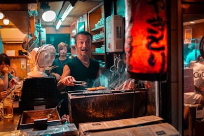 A busy and lively small Japanese restaurant kitchen with a chef grilling food over charcoal. The scene is warmly lit, and several patrons are visible enjoying their meals at the counter. The ambiance is cozy, with a red paper lantern providing ambient lighting.