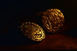 Two intricately designed gold coins rest on a dark surface. The lighting highlights the detailed carvings, featuring abstract patterns and faces, which give them an ancient and mysterious appearance.