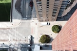 An aerial view of an urban setting with tall buildings and a street. The scene includes a white van parked near greenery, a red car parked on a dark surface, and two circular landscaped areas with trees. The ground features a combination of concrete and asphalt surfaces with various markings.