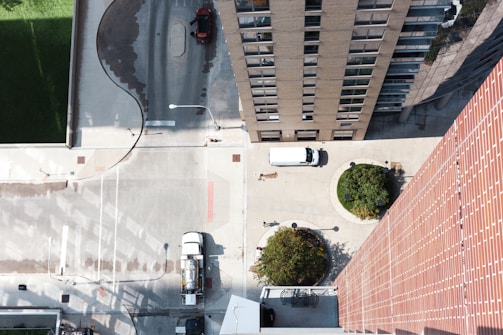 An aerial view of an urban setting with tall buildings and a street. The scene includes a white van parked near greenery, a red car parked on a dark surface, and two circular landscaped areas with trees. The ground features a combination of concrete and asphalt surfaces with various markings.
