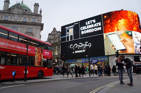 A bustling city scene featuring a prominent digital billboard displaying advertisements, including ones for a smartphone and a beverage. A classic red double-decker bus is visible to the left, and groups of people are walking and standing in the street near shops such as Gap and Boots. The architecture includes an old building with a clock and a dome.