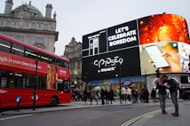 A bustling city scene featuring a prominent digital billboard displaying advertisements, including ones for a smartphone and a beverage. A classic red double-decker bus is visible to the left, and groups of people are walking and standing in the street near shops such as Gap and Boots. The architecture includes an old building with a clock and a dome.