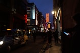 A dimly lit, bustling street features bright neon signs spelling 'Soho' mounted high on the building sides. The street is filled with pedestrians, some walking and others seemingly engaged with their phones. A traditional black taxi with illuminated taxi sign and advertisement is parked along the street, while storefronts are illuminated with various lights. The buildings have a mix of modern and classic architecture.
