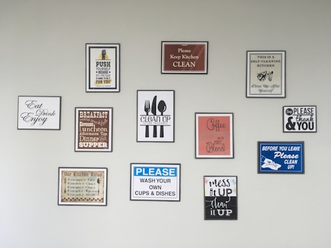 A wall features an array of framed signs with various motivational and instructional messages related to kitchen cleanliness and dining etiquette. The signs include messages such as 'Please Keep Kitchen Clean,' 'Wash Your Own Cups & Dishes,' and 'Mess It Up, Clean It Up.' There are also reminders about meal times and motivational quotes.