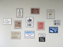 A wall features an array of framed signs with various motivational and instructional messages related to kitchen cleanliness and dining etiquette. The signs include messages such as 'Please Keep Kitchen Clean,' 'Wash Your Own Cups & Dishes,' and 'Mess It Up, Clean It Up.' There are also reminders about meal times and motivational quotes.