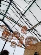 Large wicker pendant lights hang from a glass ceiling with metal supports, creating a geometric pattern. The lights are illuminated, casting a warm glow. In the bottom corner, there is partial visibility of green plants, adding a natural element to the industrial setting.
