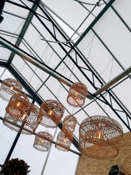 Large wicker pendant lights hang from a glass ceiling with metal supports, creating a geometric pattern. The lights are illuminated, casting a warm glow. In the bottom corner, there is partial visibility of green plants, adding a natural element to the industrial setting.
