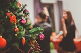 A festively decorated Christmas tree with red and pink ornaments and ribbons, positioned on the left. In the background, two people are seen blurry, enjoying a holiday gathering in a warm, inviting atmosphere.