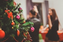 A festively decorated Christmas tree with red and pink ornaments and ribbons, positioned on the left. In the background, two people are seen blurry, enjoying a holiday gathering in a warm, inviting atmosphere.