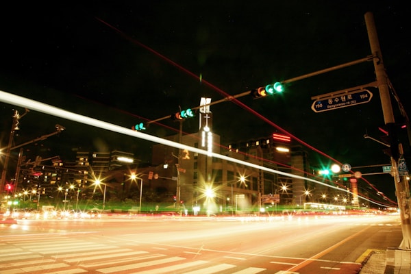 A vibrant city street intersection at night with bright streaks of light from vehicles creating dynamic trails. Traffic lights are visible, and buildings with illuminated windows line the street. The atmosphere is urban and lively.