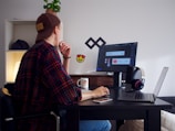 A person wearing a dark cap and a plaid shirt sits at a black desk, looking at a computer screen. The desk is cluttered with a smartphone, headphones, a laptop, and a coffee mug. A small potted plant sits on a shelf in the background alongside a lamp. The computer screen displays some graphic design work.
