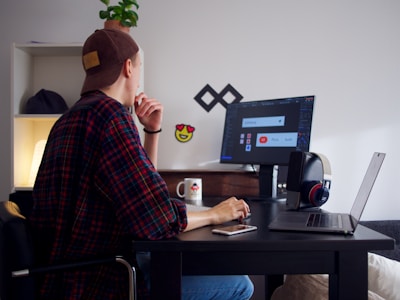 A person wearing a dark cap and a plaid shirt sits at a black desk, looking at a computer screen. The desk is cluttered with a smartphone, headphones, a laptop, and a coffee mug. A small potted plant sits on a shelf in the background alongside a lamp. The computer screen displays some graphic design work.