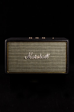 A vintage-style speaker with the brand name prominently displayed in white script against a woven fabric grille. The speaker casing has a textured black finish and gold control knobs on top, set against a black background.