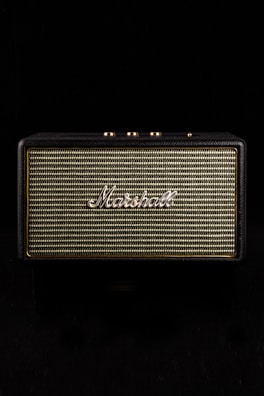 A vintage-style speaker with the brand name prominently displayed in white script against a woven fabric grille. The speaker casing has a textured black finish and gold control knobs on top, set against a black background.