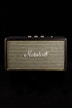 A vintage-style speaker with the brand name prominently displayed in white script against a woven fabric grille. The speaker casing has a textured black finish and gold control knobs on top, set against a black background.