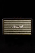 A vintage-style speaker with the brand name prominently displayed in white script against a woven fabric grille. The speaker casing has a textured black finish and gold control knobs on top, set against a black background.