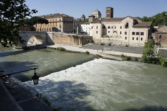 A scenic river flows under a stone arch bridge, surrounded by historic buildings. The architecture features classic European design with brickwork and terra-cotta rooftops. Trees frame the scene, adding lush greenery, while the sunlight casts sharp shadows, giving a warm and inviting atmosphere.