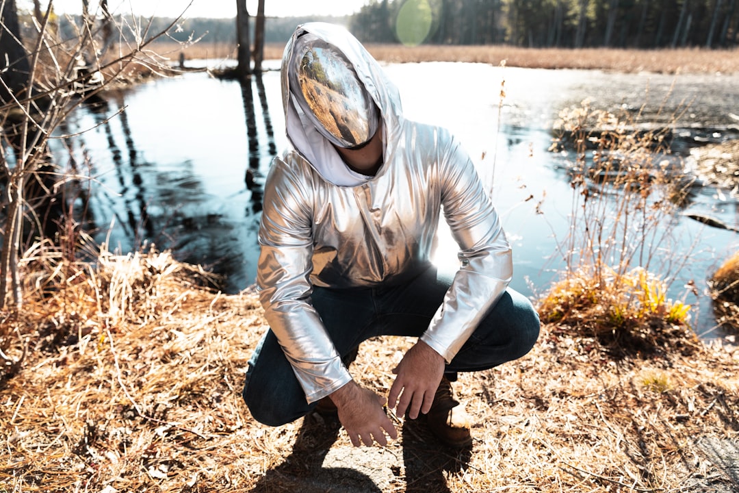 man in silver jacket kneeling on grass near pond during daytime