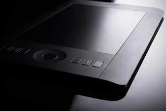 A sleek, modern graphics tablet with a dark surface and multiple buttons along the edges. The tablet is positioned against a balanced contrast of light and shadow, highlighting its refined and professional design.