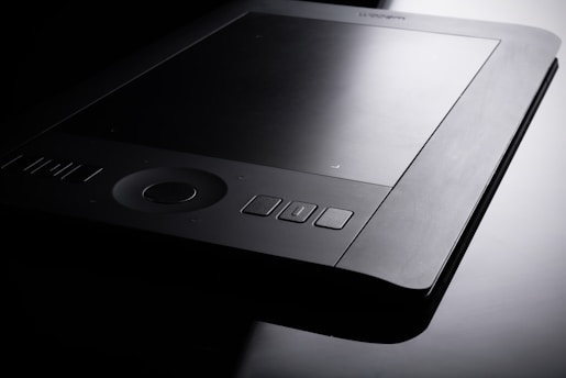 A sleek, modern graphics tablet with a dark surface and multiple buttons along the edges. The tablet is positioned against a balanced contrast of light and shadow, highlighting its refined and professional design.