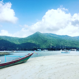 A serene beach setting with a traditional wooden boat resting on sandy shores. In the background, lush green hills rise under a blue sky scattered with white clouds. The scene includes gentle waves lapping against the shore and a few distant boats on the water.
