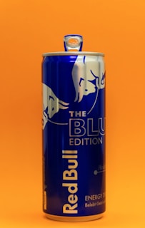 A blue can of Red Bull energy drink stands against a bright orange background. The can features the Red Bull logo and 'The Blue Edition' label prominently displayed.