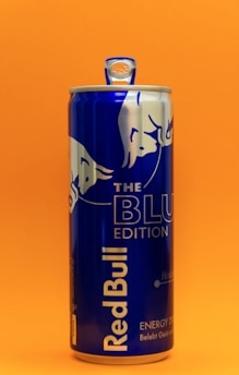 A blue can of Red Bull energy drink stands against a bright orange background. The can features the Red Bull logo and 'The Blue Edition' label prominently displayed.