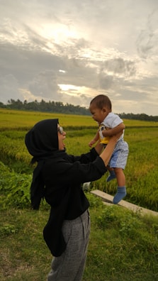 A woman wearing a black hijab and sunglasses is joyfully lifting a baby in the air outdoors. They are surrounded by lush green fields under a cloudy sky with the sun peeking through.