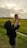 A woman wearing a black hijab and gray pants holds a smiling baby high in the air against a backdrop of lush green fields and a cloudy sky at sunset.