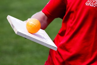 A person wearing a red shirt is balancing an orange plastic ball on a white wooden board. The background features a grassy area.