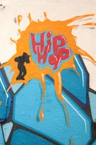 A vibrant graffiti artwork features the words 'Hip Hop' in bold, stylized lettering. The text is highlighted with an orange paint splash and is accompanied by a small silhouette of a person, dynamically depicted in motion. The background consists of blue geometric shapes with black outlining.