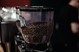 A close-up view of a transparent coffee bean grinder filled with roasted coffee beans. The grinder is labeled and appears to be part of a series of similar equipment arranged in a row. The background is softly blurred, focusing the attention on the grinder and its contents.