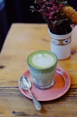 A glass of matcha latte with latte art on top, presented on a pink saucer with a spoon on the side. The scene also includes a bouquet of dried flowers in a decorative white vase, placed on a wooden table.