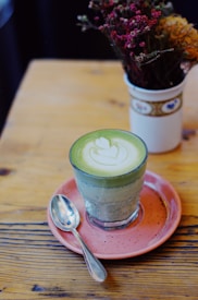A glass of matcha latte with latte art on top, presented on a pink saucer with a spoon on the side. The scene also includes a bouquet of dried flowers in a decorative white vase, placed on a wooden table.
