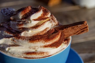 A close-up view of a swirl of whipped cream topped with cocoa powder and a chocolate flake placed in a blue cup. The texture is rich and creamy, with the chocolate flake adding a crunchy element.
