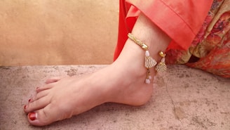 A close-up view of a foot adorned with a detailed, decorative anklet featuring intricate gold and white designs. The toenails are painted red, and the foot is resting on a textured, stone-like surface. An orange garment is partially visible.