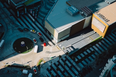 The aerial view captures an intersection with a circular roundabout featuring a small green area with a structure in the center. Several vehicles, including cars and a white van, are driving around it. A large building with the name 'MERAAS' on the facade is adjacent to the road, with a patterned pavement visible. The lighting creates sharp shadows, enhancing contrast between the sunlit and shaded areas. The scene appears to be in an urban environment with organized lanes and pedestrian areas.