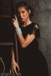 A woman with styled brown hair is leaning against a wooden ladder. She is wearing a black dress with lace detailing and adorned with multiple silver bracelets on her wrist. The surroundings have a rustic, textured wall and some green foliage.