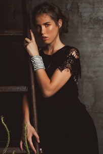 A woman with styled brown hair is leaning against a wooden ladder. She is wearing a black dress with lace detailing and adorned with multiple silver bracelets on her wrist. The surroundings have a rustic, textured wall and some green foliage.