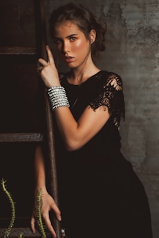 A woman with styled brown hair is leaning against a wooden ladder. She is wearing a black dress with lace detailing and adorned with multiple silver bracelets on her wrist. The surroundings have a rustic, textured wall and some green foliage.
