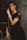A woman with styled brown hair is leaning against a wooden ladder. She is wearing a black dress with lace detailing and adorned with multiple silver bracelets on her wrist. The surroundings have a rustic, textured wall and some green foliage.