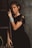 A woman with styled brown hair is leaning against a wooden ladder. She is wearing a black dress with lace detailing and adorned with multiple silver bracelets on her wrist. The surroundings have a rustic, textured wall and some green foliage.